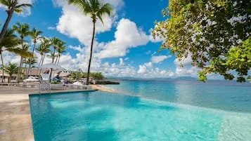 A luxurious swimming pool overlooking the ocean with palm trees, lounge chairs, and a partly cloudy sky.