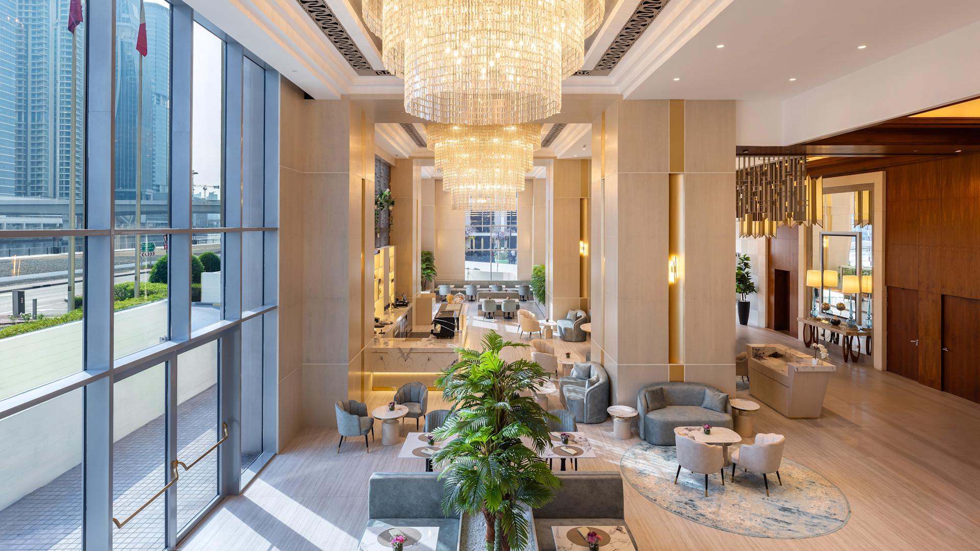Luxurious hotel lobby with high ceilings, chandeliers, large glass windows, and comfortable seating areas with chairs and tables, decorated with plants and modern lighting.