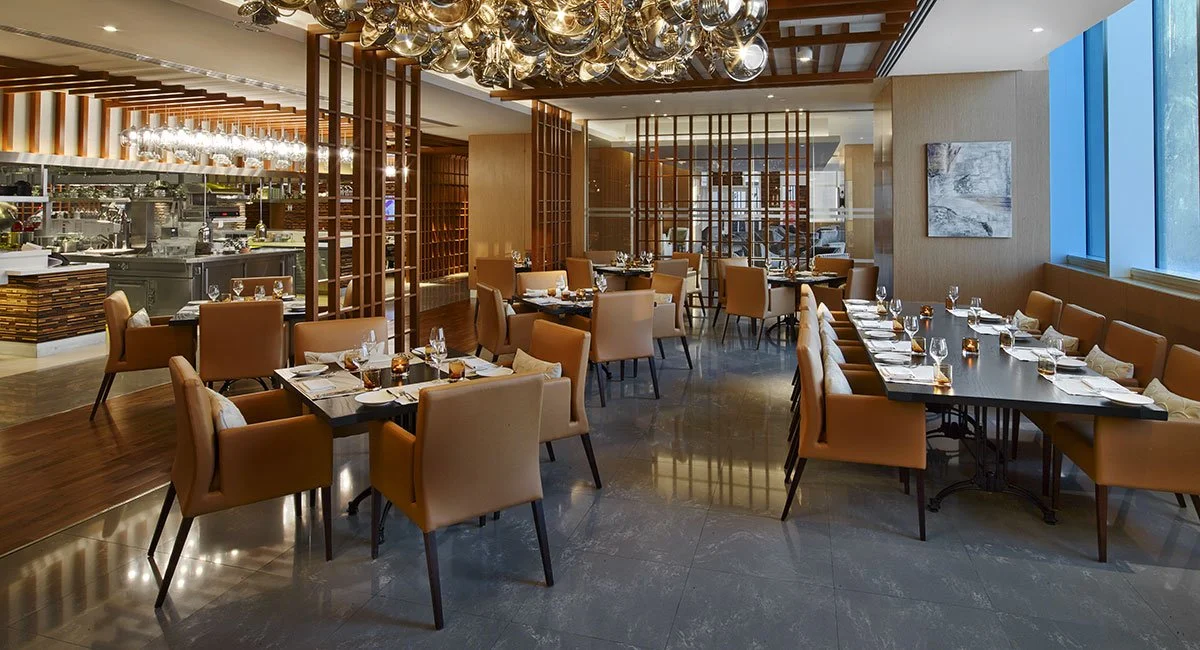 Elegant restaurant dining area with tables set with wine glasses, napkins, and silverware, tan upholstered chairs, modern lighting fixtures, and a view of the kitchen area in the background.