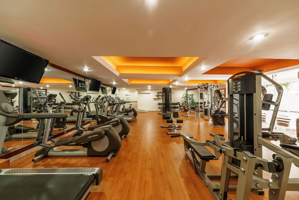 Empty gym with various exercise equipment, treadmill, elliptical, and weight machines on hardwood floors, illuminated ceiling lights, and some wall-mounted TV screens.
