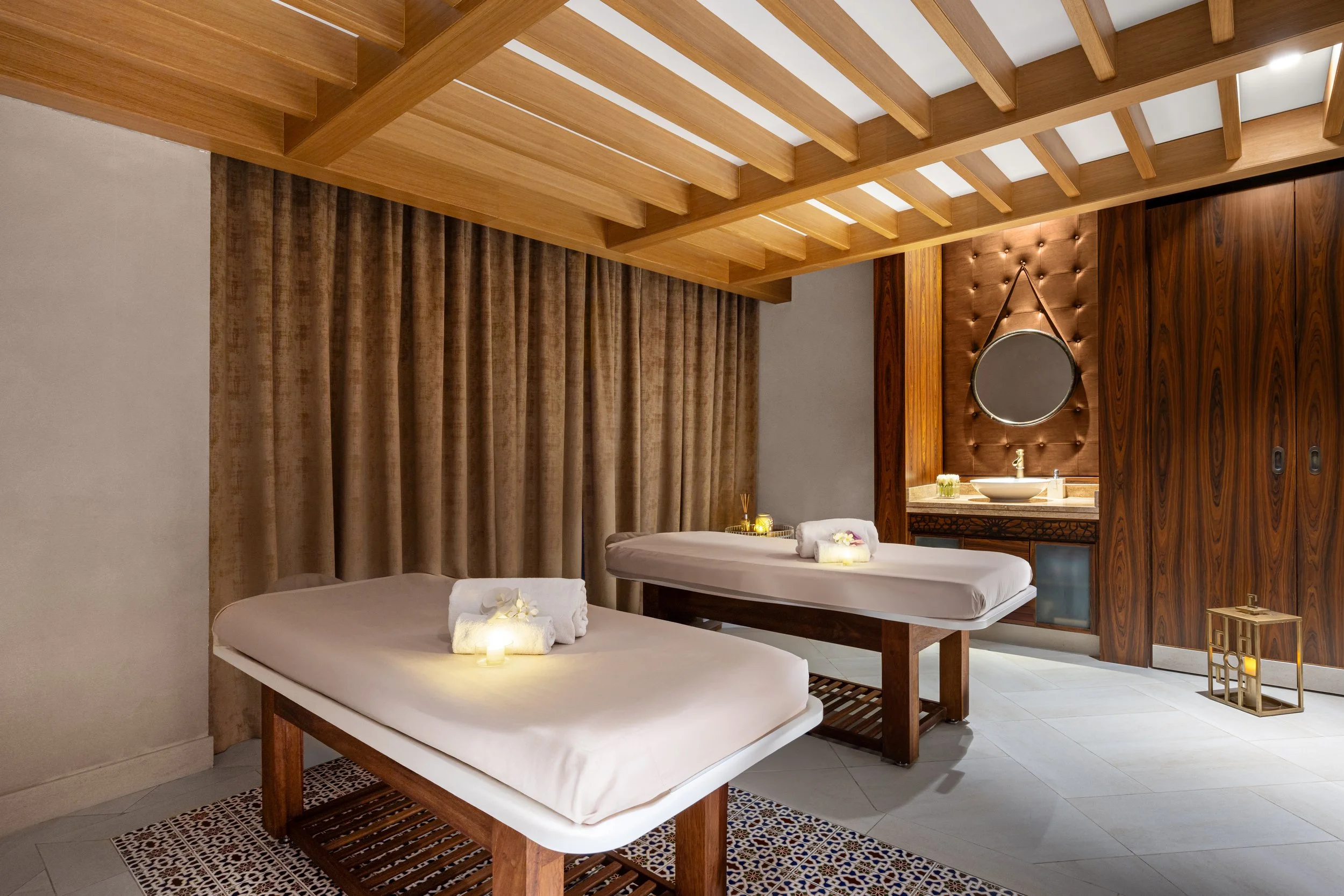 Two massage beds with rolled towels and flowers in a spa room with wooden ceiling beams, beige curtains, and a bathroom mirror.