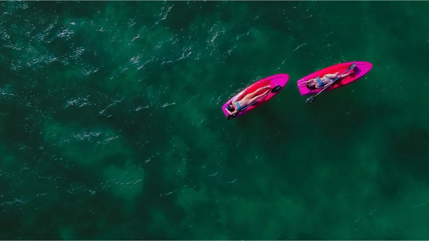Two people lying on pink paddleboards in a green ocean, relaxing and paddling.