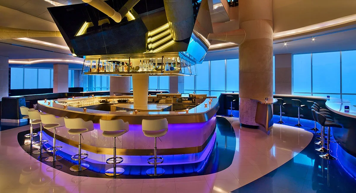 Upscale bar with a curved counter, white barstools, and blue accent lighting, overlooking large windows with a city view at night.