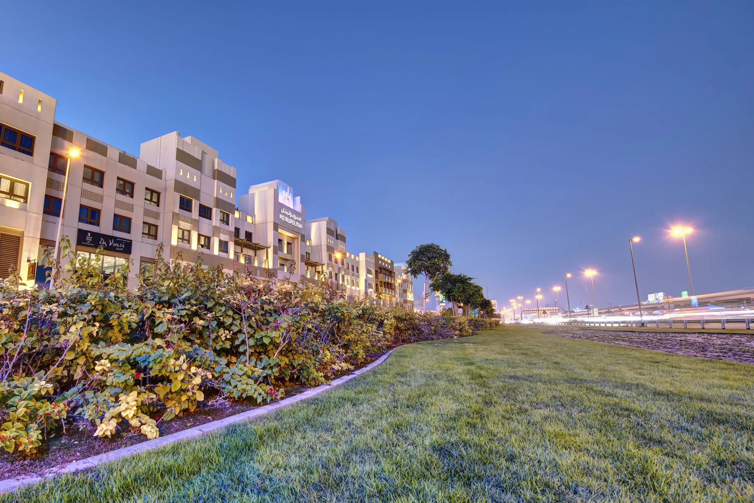 Cityscape at dusk with modern white buildings, street lights, green grass, and a clear blue sky.