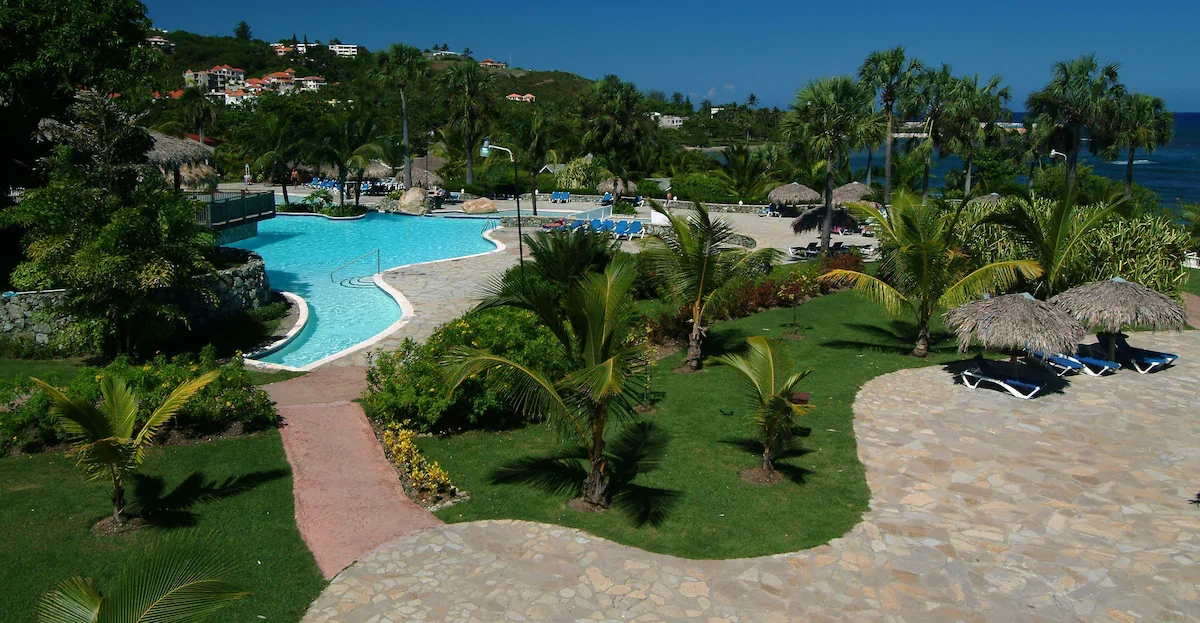 Resort swimming pool area surrounded by palm trees, with lounge chairs, parasols, and a stone pathway, overlooking houses on a hillside and the ocean in the background.