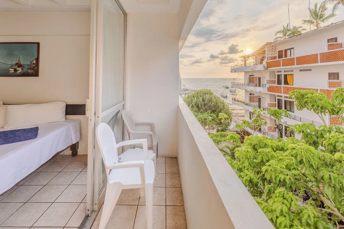 View from a balcony overlooking a hotel or apartment complex with a sea and sunset in the distance, and trees in the foreground.