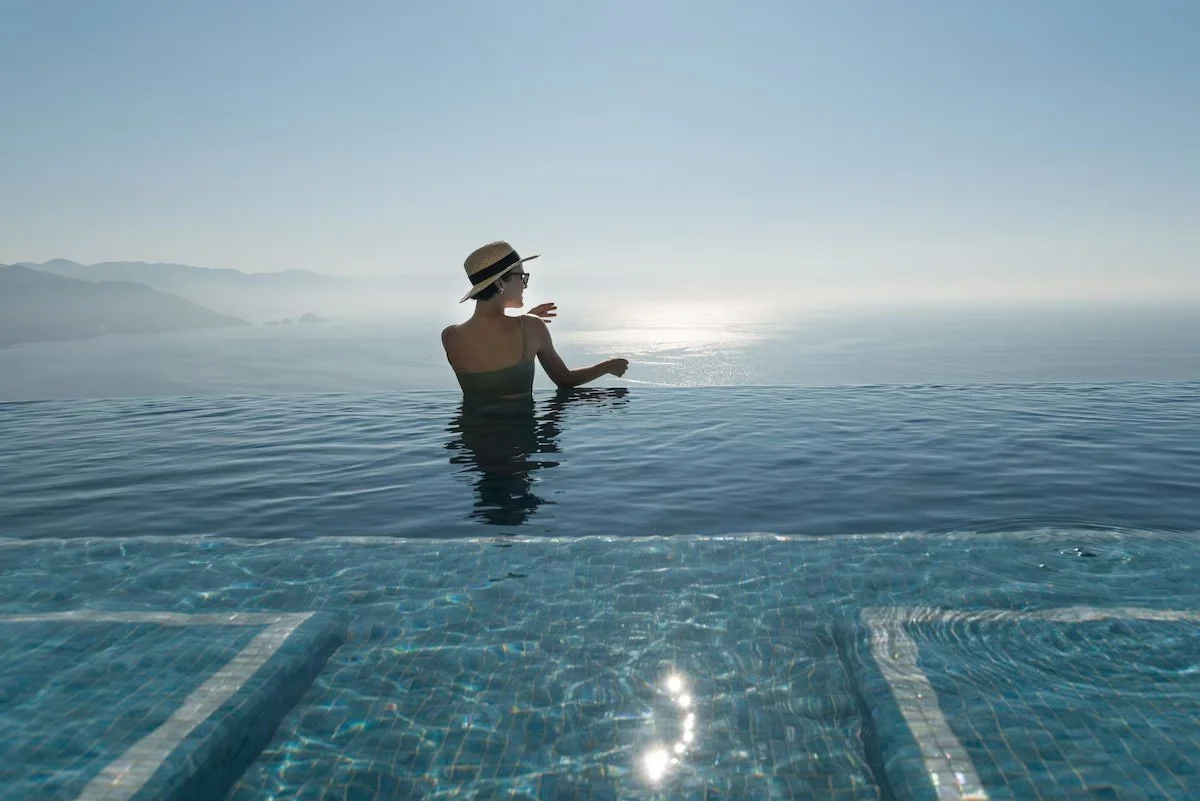 A woman in a black swimsuit and straw hat standing in an infinity pool, facing the ocean with mountains in the background, during sunset or sunrise.