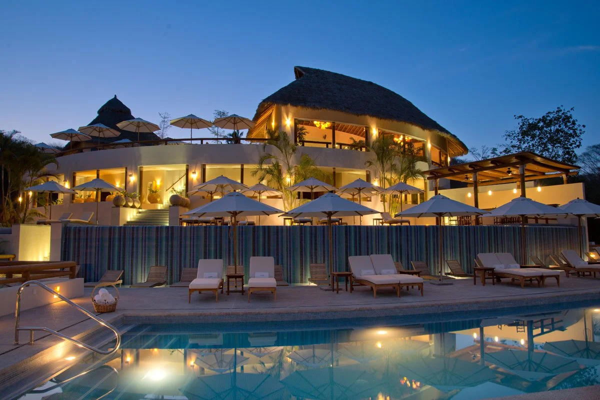 Night view of a luxurious outdoor pool area with lounge chairs, closed umbrellas, and a multi-level building with thatched roof, warm lighting, and patio seating, set against a darkening sky.