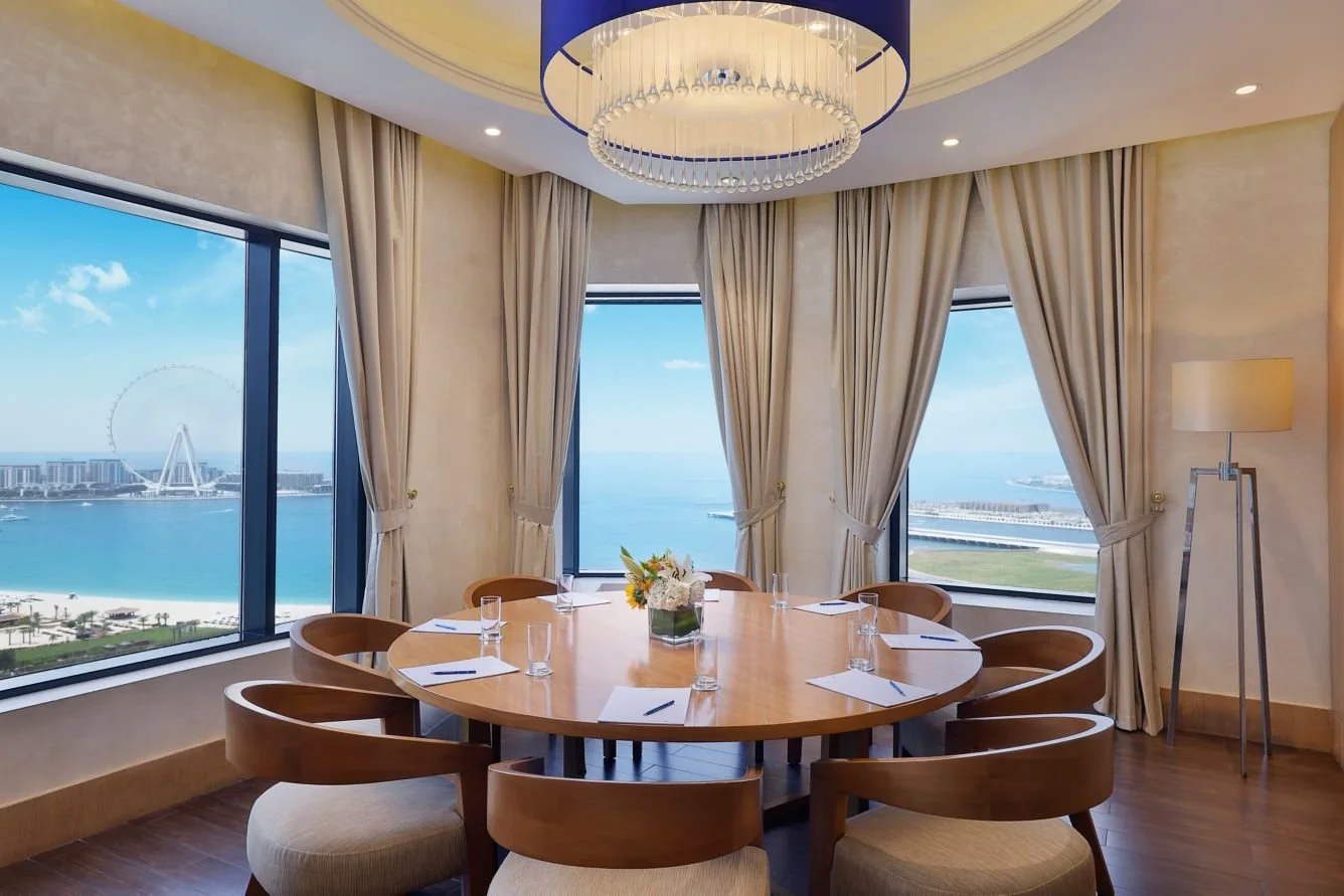 Elegant conference room with a round wooden table set with notepads and pens, surrounded by eight chairs, large windows showing a waterfront cityscape with a Ferris wheel, and a view of the water and distant buildings, with beige curtains and a moder