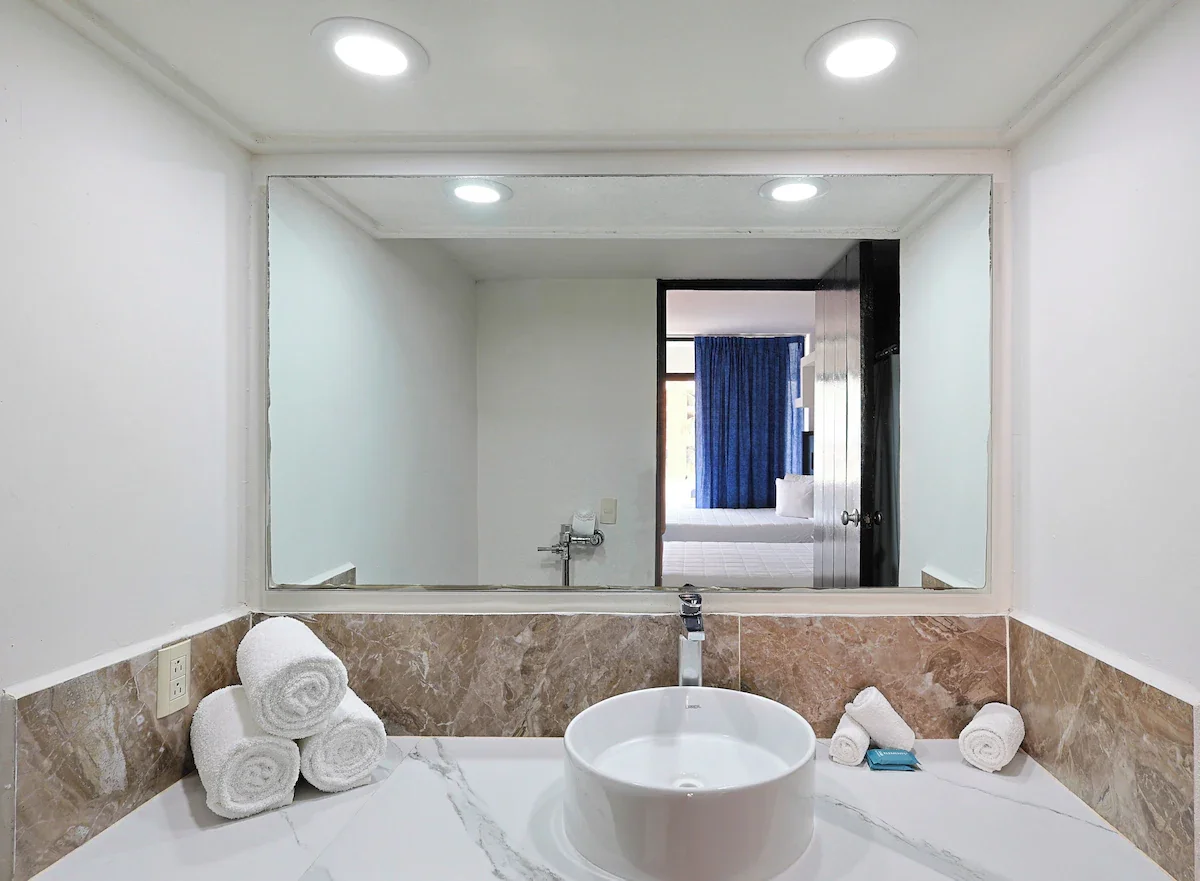Bathroom with a large mirror, white marble countertop, stacked white towels, a round vessel sink, and a wall outlet, with a bedroom visible through the mirror reflection.