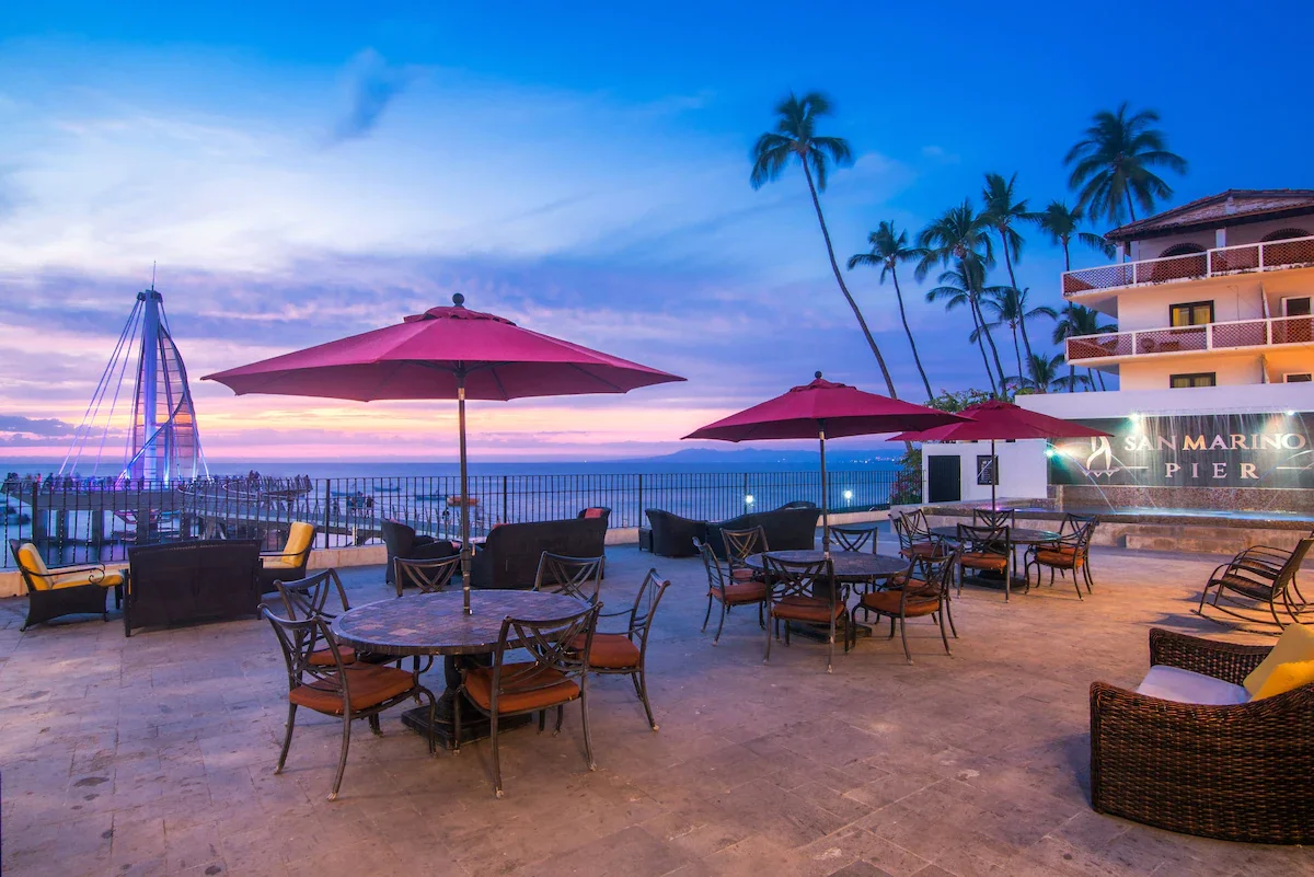 Outdoor patio with tables and chairs under pink umbrellas, overlooking the ocean during sunset, with palm trees and a building labeled San Marino Pier.