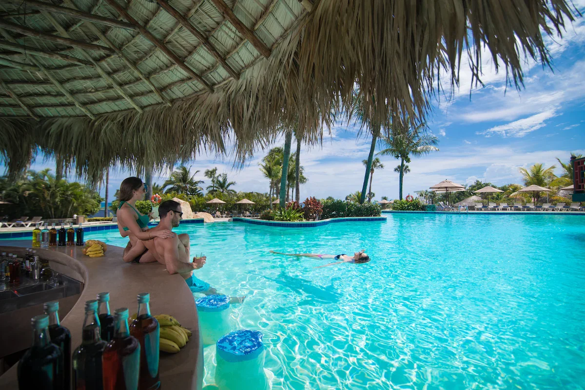 A tropical poolside scene with a thatched roof over the bar area, two people sitting on the bar's edge, and a woman floating on her back in the pool. Palm trees and lounge chairs with umbrellas are in the background, under a partly cloudy sky.