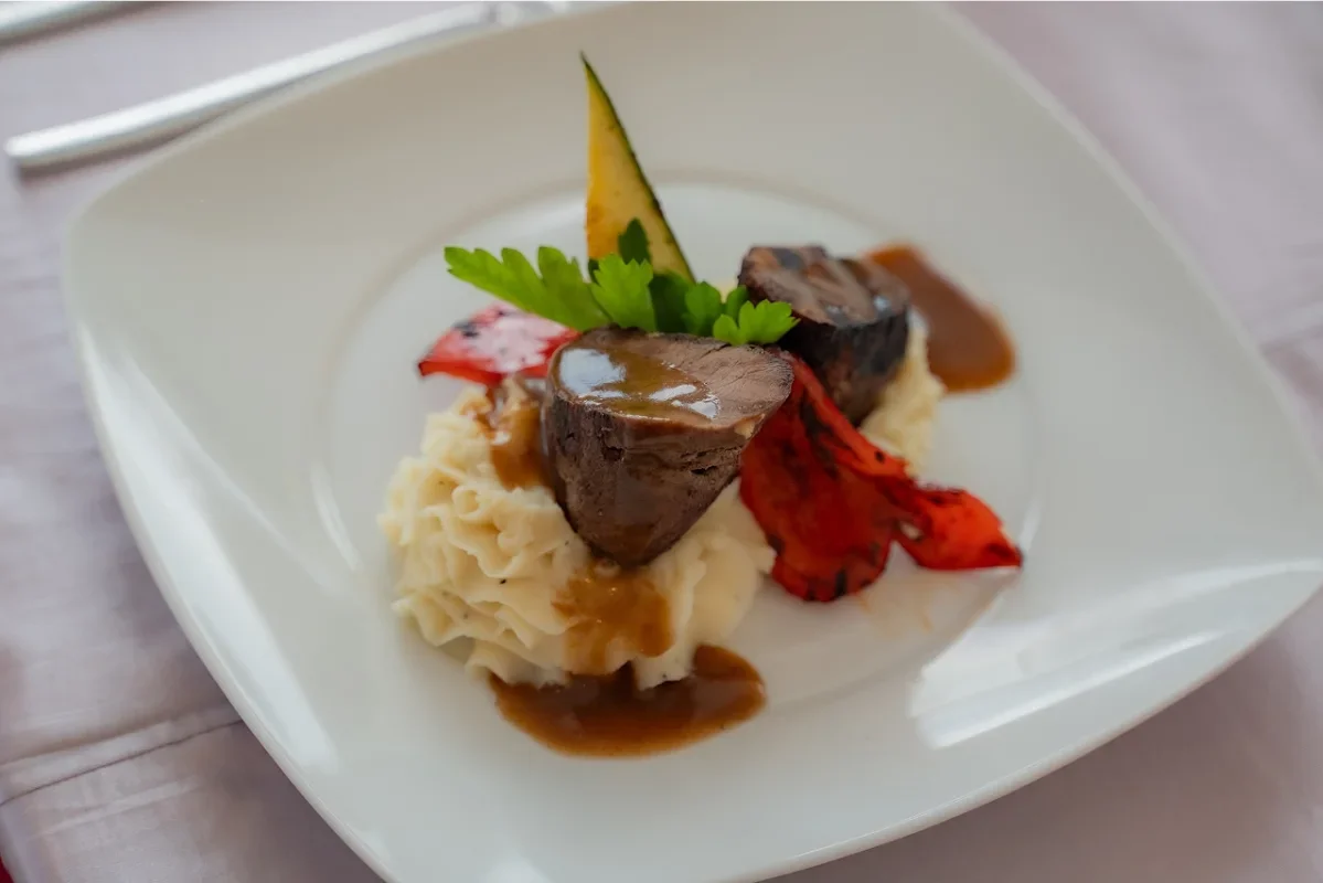 A plated gourmet dish with mashed potatoes, two pieces of steak topped with glaze, red and yellow bell peppers, garnished with leafy herbs and a zucchini slice, on a white plate.