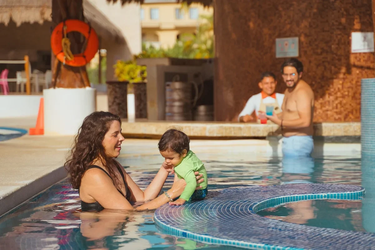 A woman in a swimsuit holding a baby in a swimming pool, with two men in the background enjoying drinks at the poolside bar.