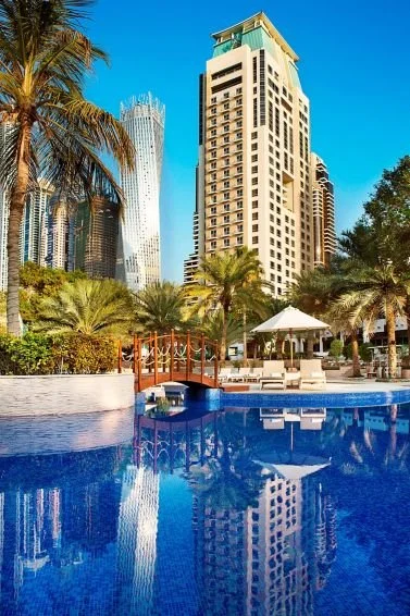 City skyline with tall buildings, palm trees, pool with reflections, and outdoor seating area with umbrellas.