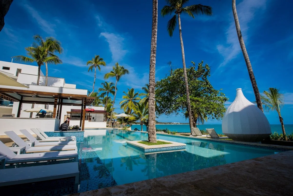A tropical resort swimming pool area with palm trees, lounge chairs, and a white cabana, overlooking the ocean under a clear blue sky.