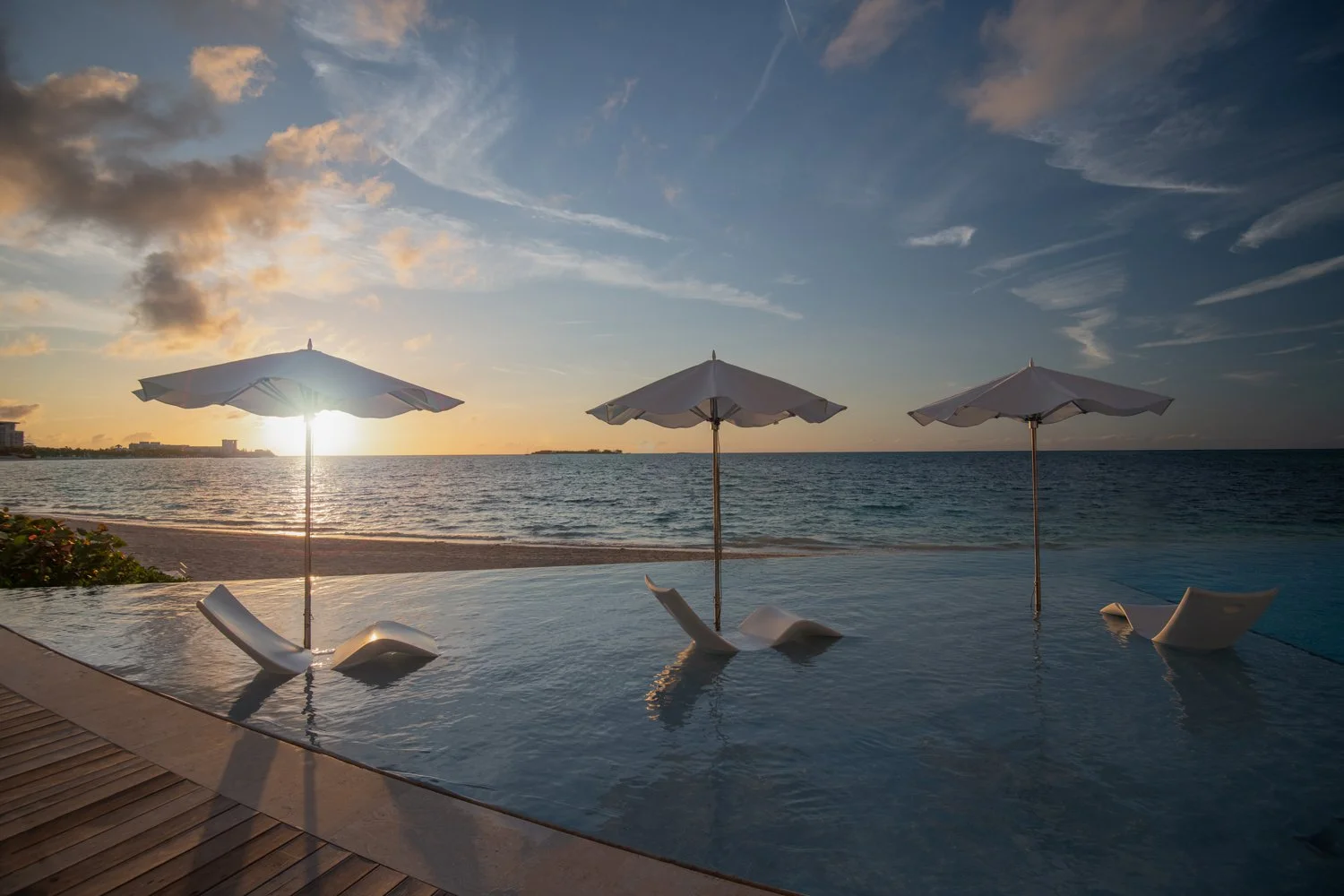 Sunset view of a beachside pool with three white umbrellas and lounge chairs, overlooking the ocean under a partly cloudy sky.