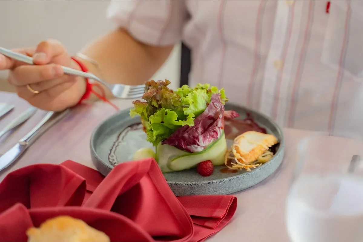 A person holding a fork about to eat a modern salad with leafy greens, rolled cucumber, and a small piece of bread or pastry on a gray plate. A red cloth napkin is on the table.