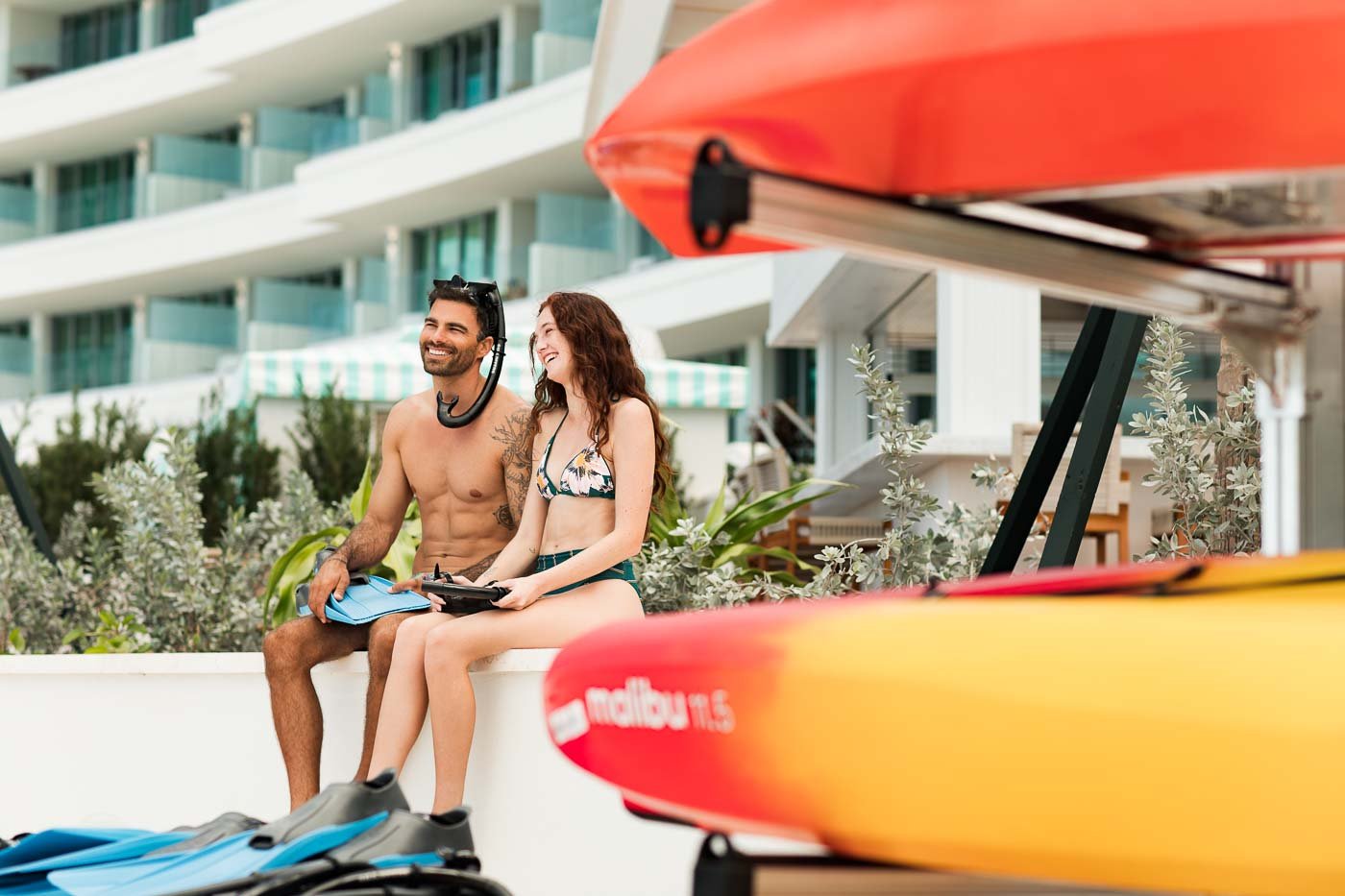 A young couple sitting on a white ledge near the beach, smiling and enjoying the sunshine, with kayaks and snorkeling gear in front of them, and a modern building with balconies in the background.