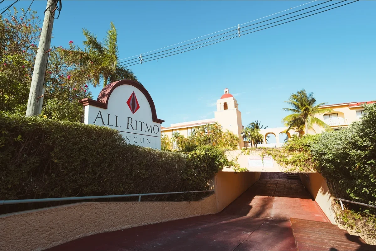 Entrance to All Ritmo Cancun with a sign, greenery, palm trees, and a building with arches and a bell tower in the background, under a clear blue sky.