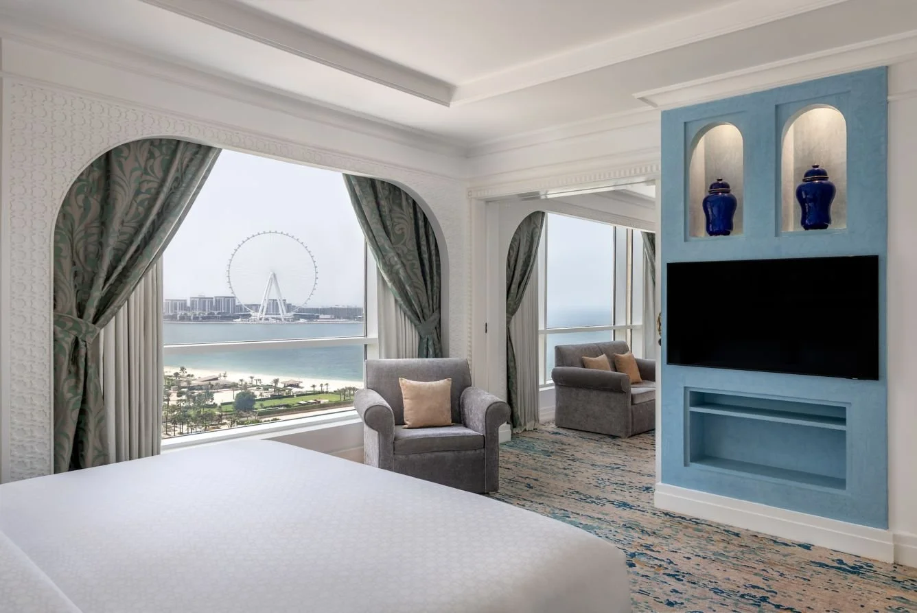 A hotel room with large windows showing a view of a Ferris wheel, water, and cityscape, furnished with gray armchairs, beige pillows, curtains, and a blue entertainment center with decorative vases.