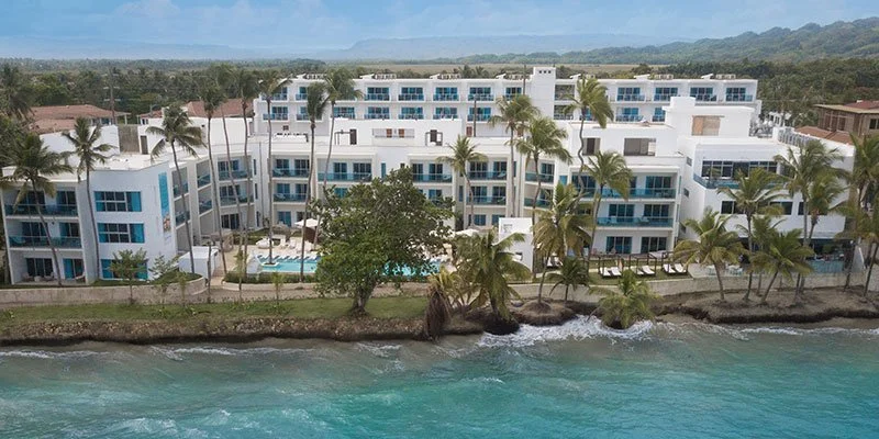 Aerial view of a modern beachfront condominium complex with white buildings, palm trees, and a swimming pool, located along the shoreline of the ocean with clear blue water.