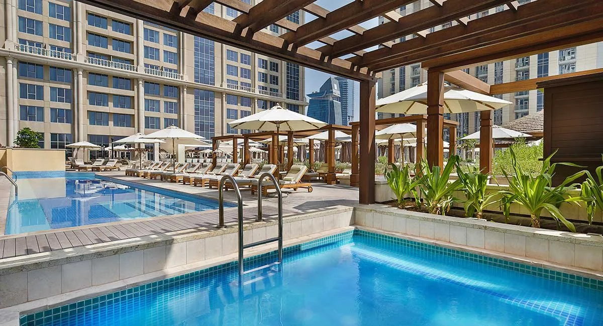 Swimming pool area at a modern high-rise apartment complex with lounge chairs, umbrellas, and surrounding green plants.