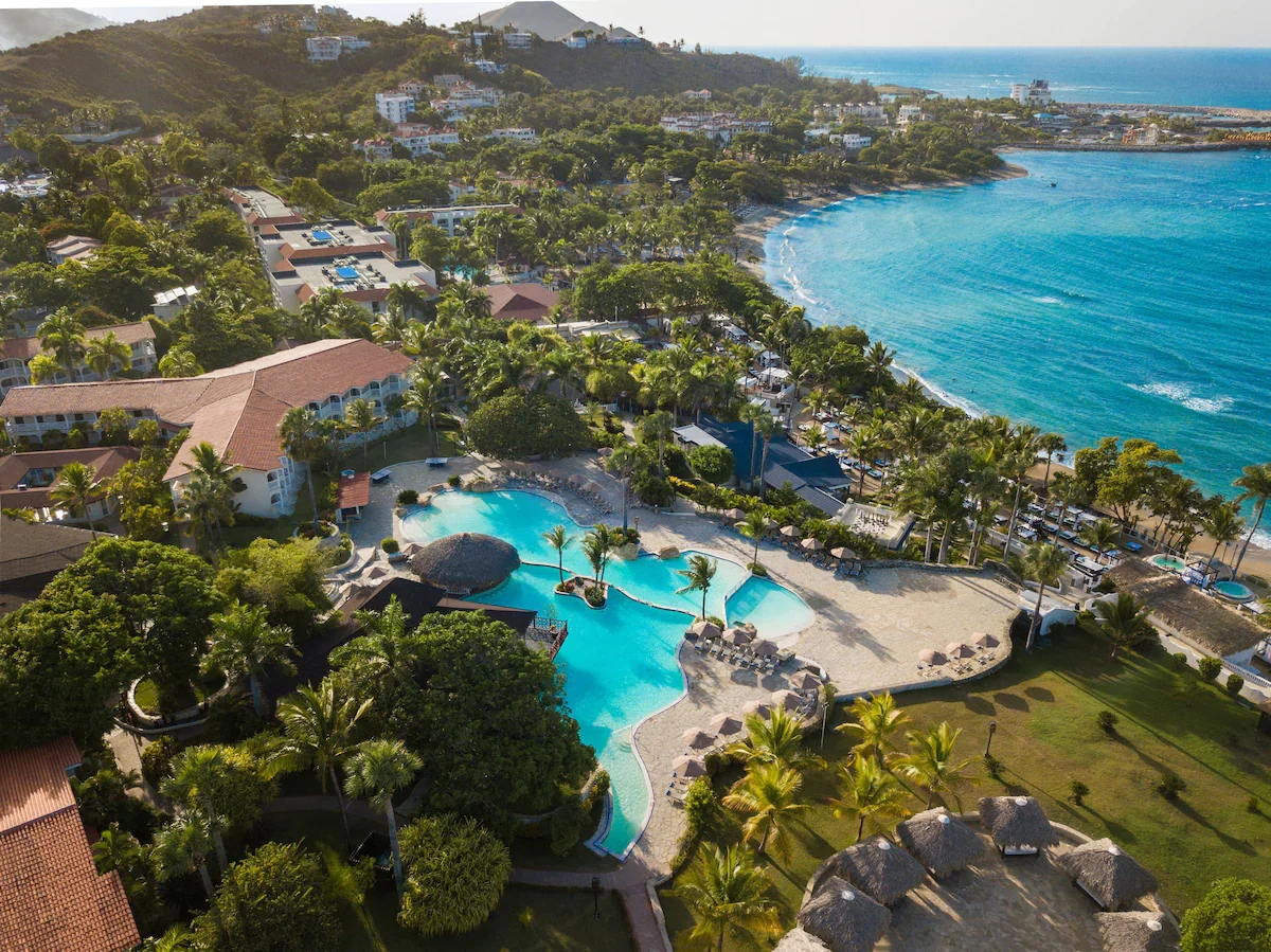 Aerial view of a tropical beach resort with a large pool surrounded by palm trees, lush greenery, and beachfront buildings along the ocean shoreline.
