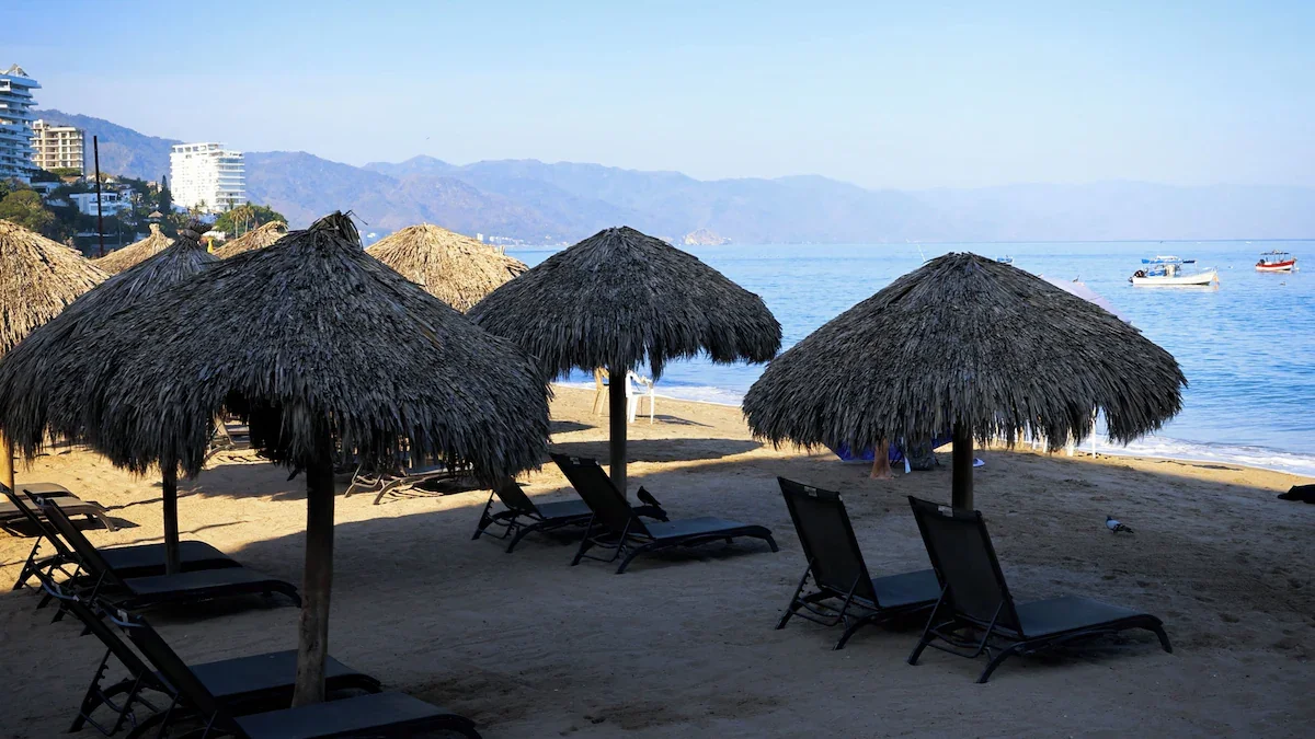 Beach with straw umbrellas and lounge chairs, calm water with boats, mountains in the background.