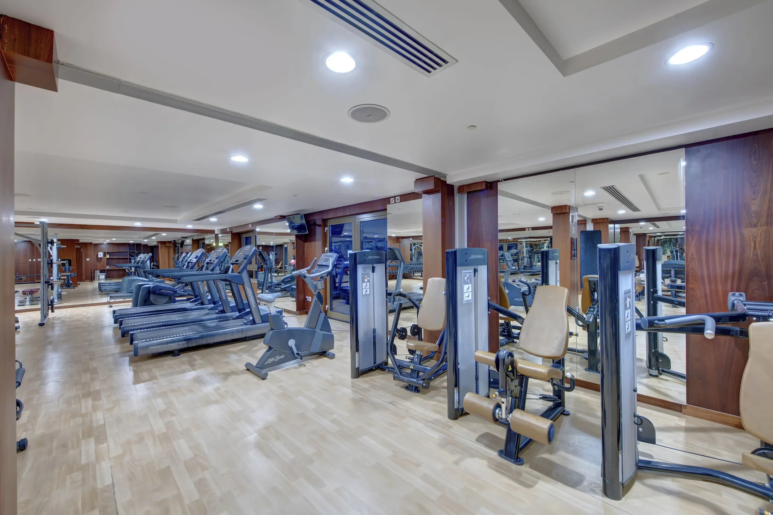 Fitness gym with treadmills, elliptical machines, and weight training equipment, featuring wood paneling, large mirrors, and bright ceiling lights.