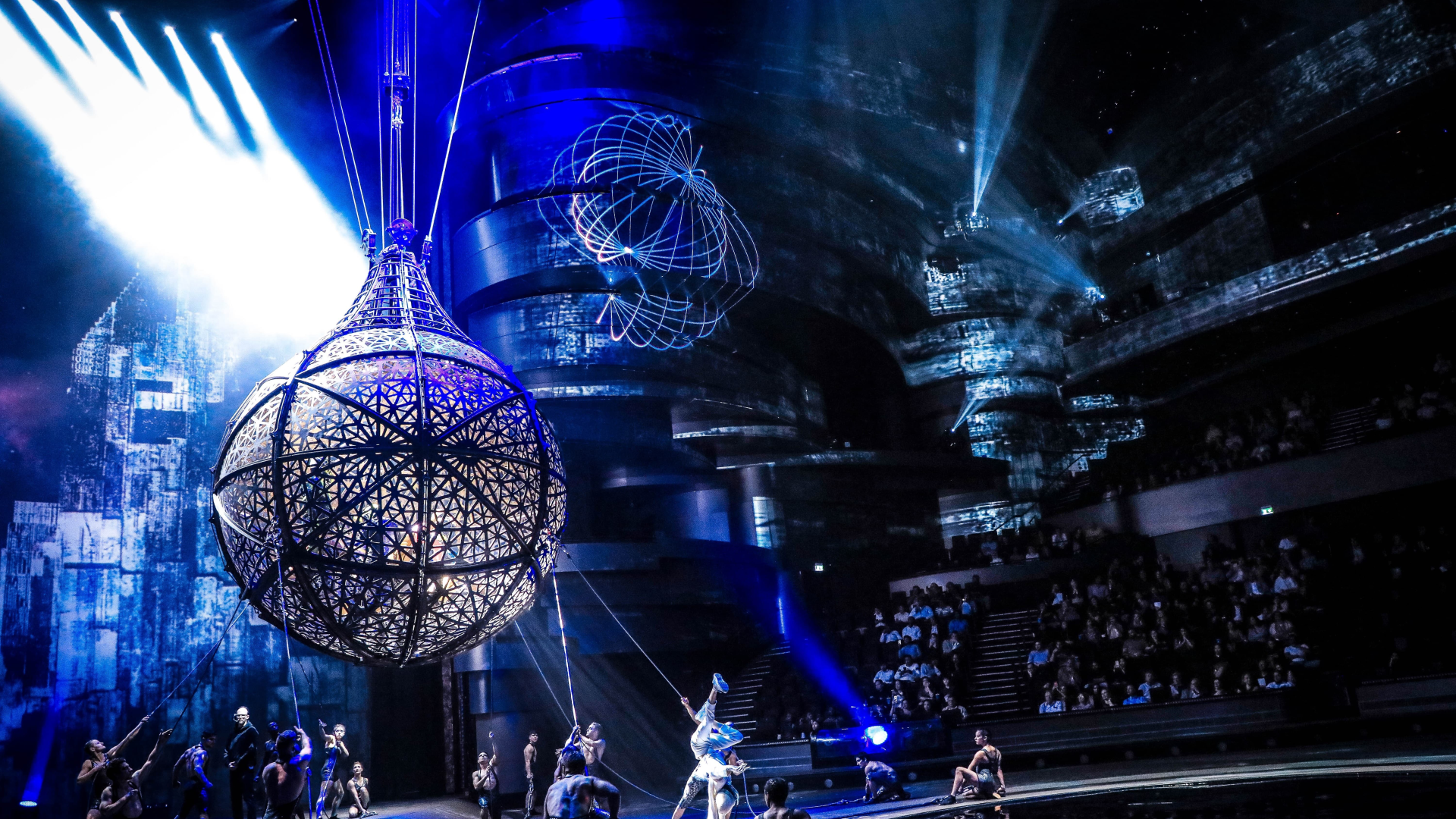 Aerial performers and acrobats in a circus show with bright blue and white stage lighting, featuring large globe-shaped props and LED globe graphics projected on the background walls, with an audience watching from tiered seating.