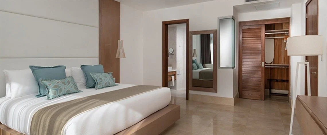 Hotel room with a large bed, blue and patterned pillows, wooden accents, a closet, a mirror, and an adjoining bathroom.