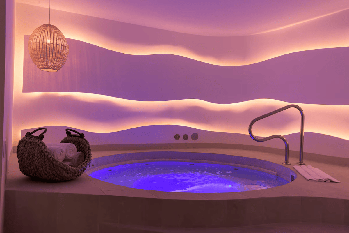 Indoor spa room with purple lighting, a small round hot tub, a woven basket with rolled towels, and wave-patterned wall panels.