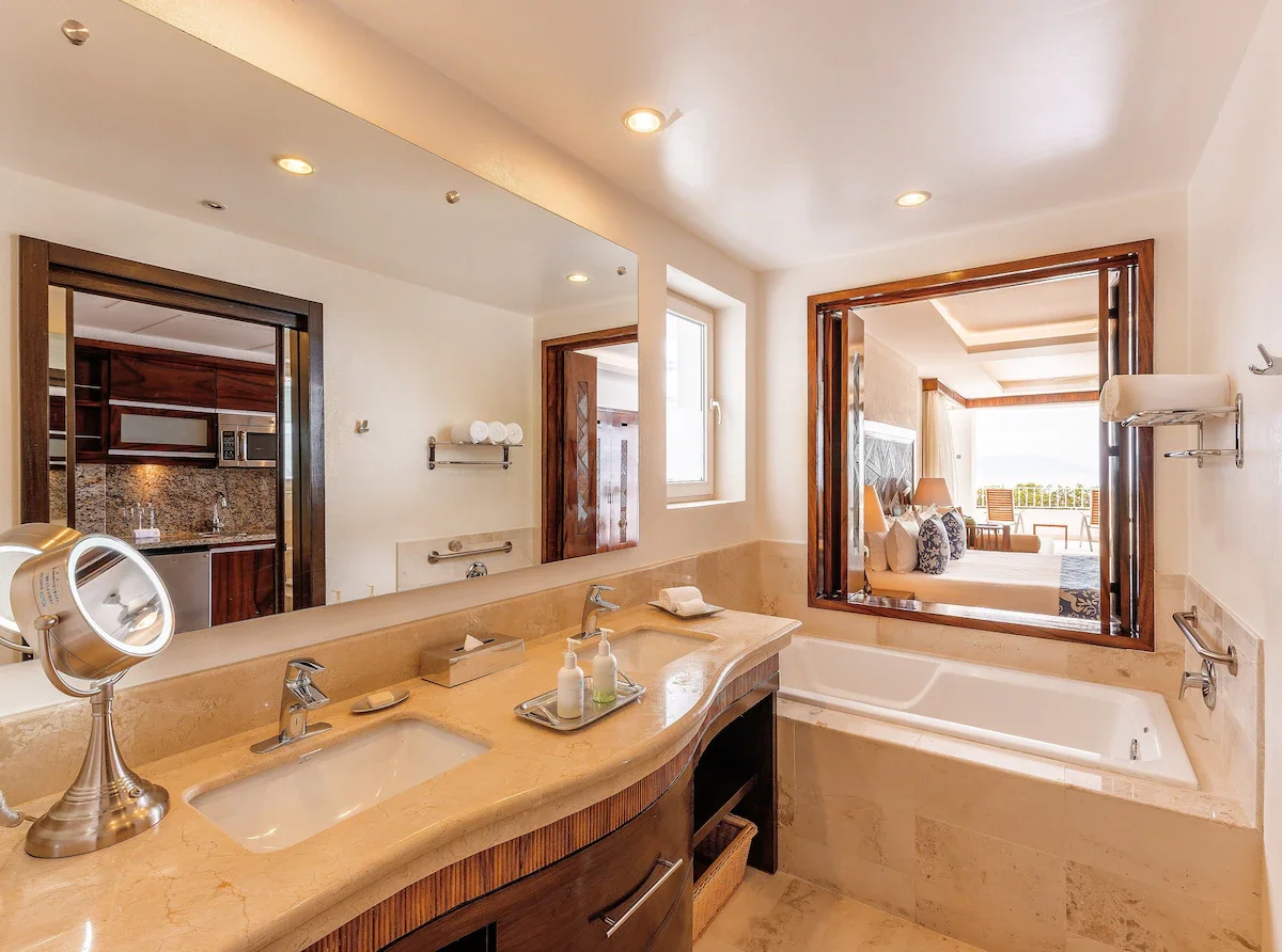 Hotel bathroom with dual sinks, marble countertop, bathtub, large mirror, window, and view into a bedroom with bed and seating area.