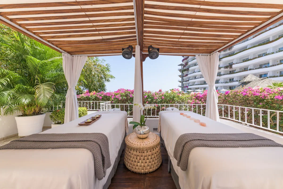 Two massage tables with white sheets and gray towels in an outdoor spa setting under a wooden canopy, overlooking a balcony with pink flowering bushes and a modern building in the background.