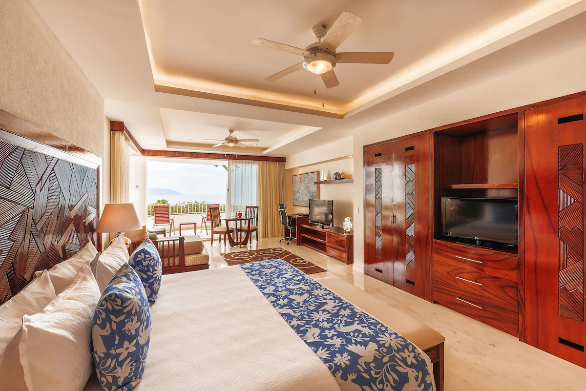 A spacious hotel room with a large bed, decorative blue and white pillows, a bedside lamp, a sitting area near a large window with patio furniture, a wooden entertainment unit with a flat-screen TV, and a wooden wardrobe. There is a ceiling fan and r