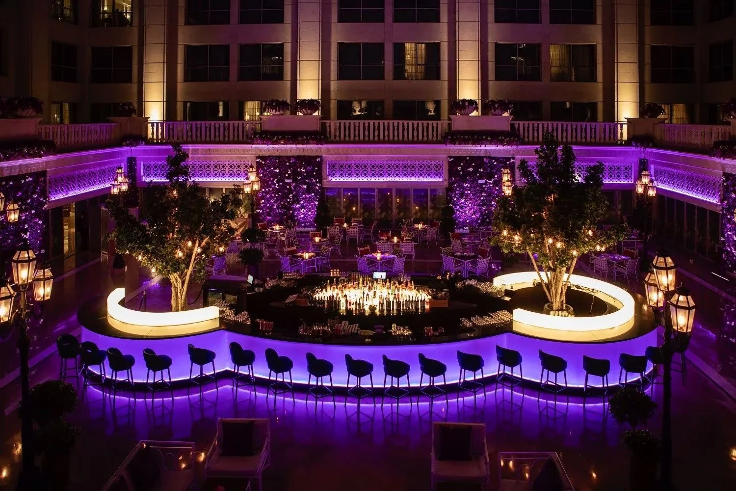 Night view of a luxurious hotel lobby or bar area illuminated with purple lighting, featuring a central bar with bar stools, trees, and small tables with chairs, surrounded by balconies and large windows.