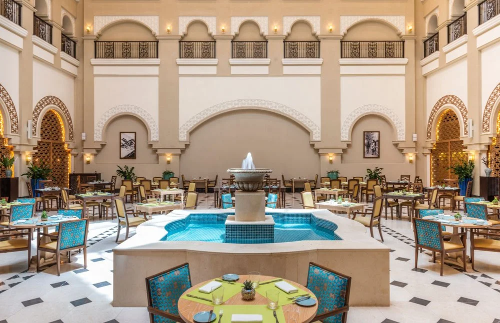 Interior of an upscale hotel or banquet hall with a central fountain, multiple dining tables with chairs, and decorative arches and Scottish-style wall decor.