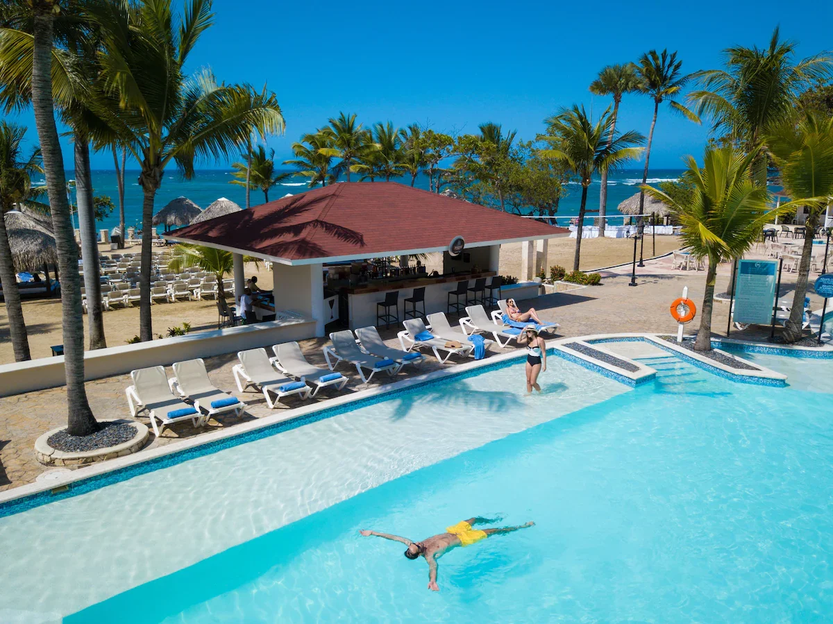 Resort pool area with lounge chairs and a bar, surrounded by palm trees, overlooking the ocean. People are relaxing in the pool and on the poolside chairs, with thatched roof structures and blue skies.