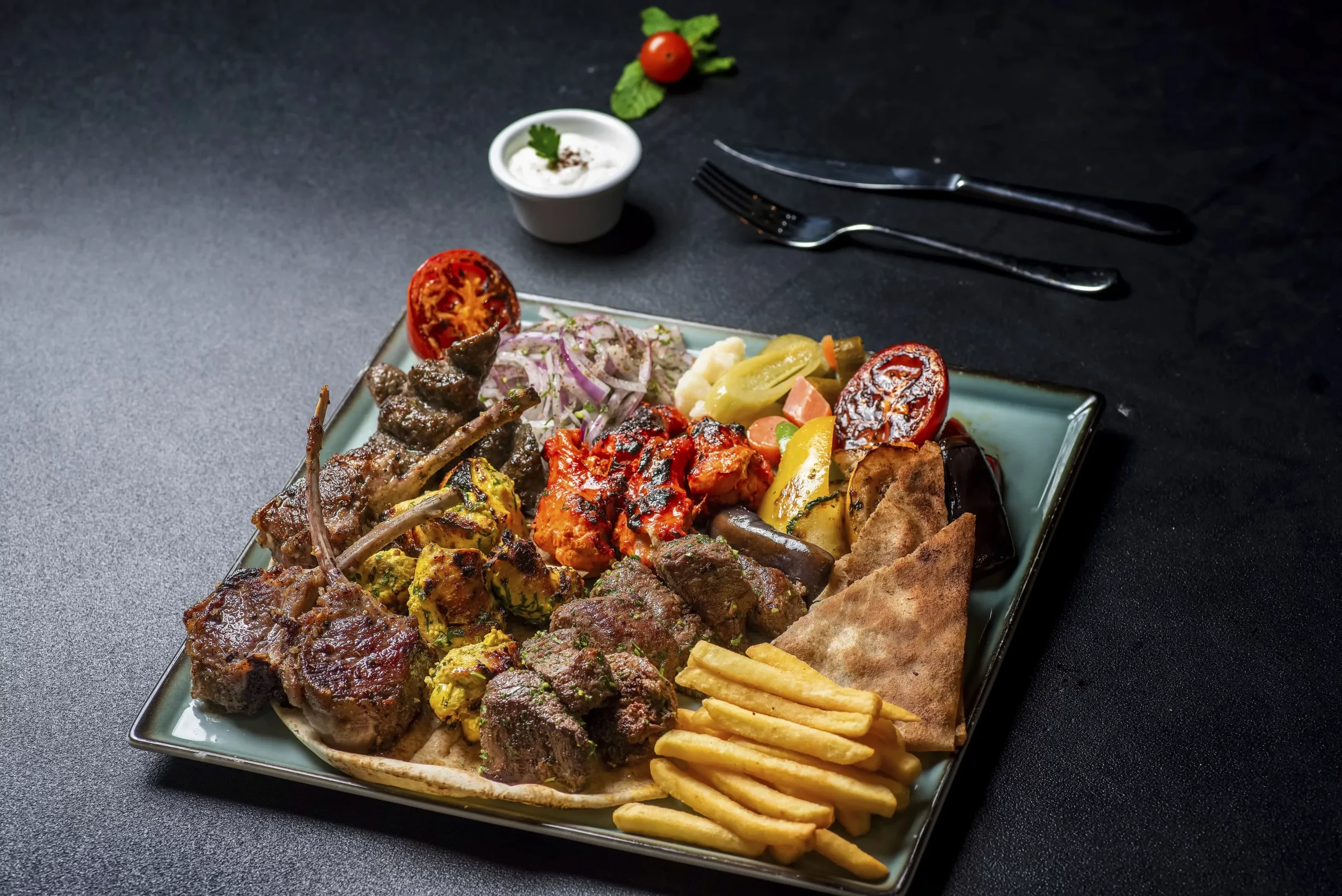 A plate of mixed grilled meats, vegetables, and fries with a side of tomato and onion salad, served on a rectangular green plate.