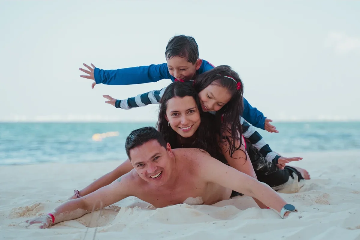 A family of four playing on the beach, stacking on top of each other, with the ocean in the background.