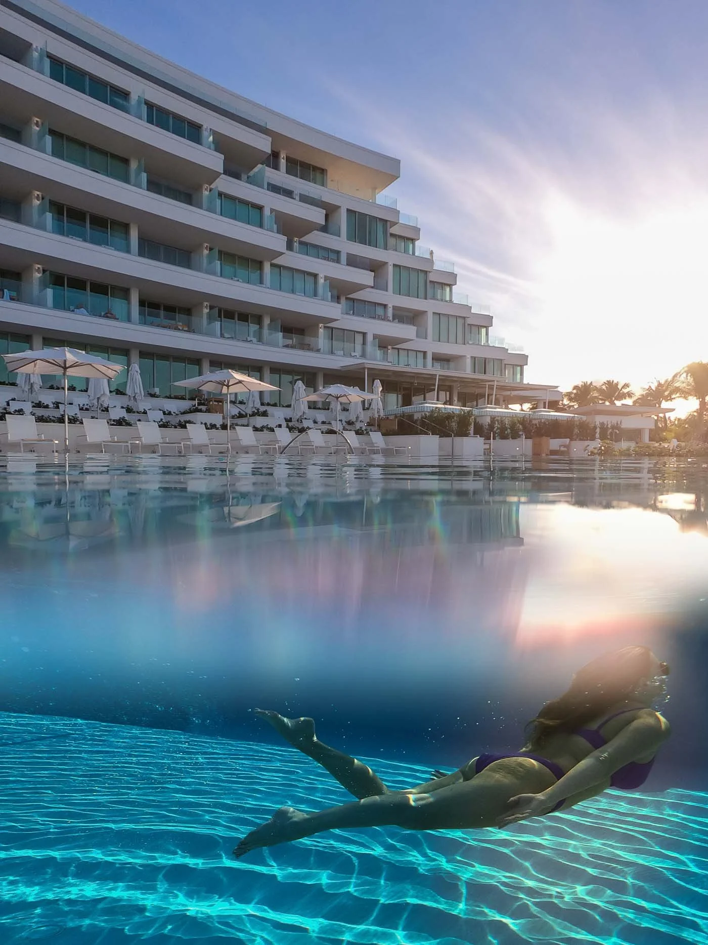 Woman swimming in an outdoor pool with a modern building and palm trees in the background, during sunset.