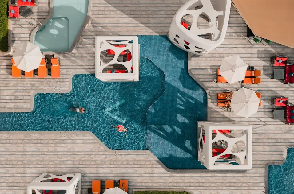 An aerial view of a swimming pool area with people swimming, surrounded by orange and white lounge chairs, umbrellas, and modern white cabanas on a wooden deck.