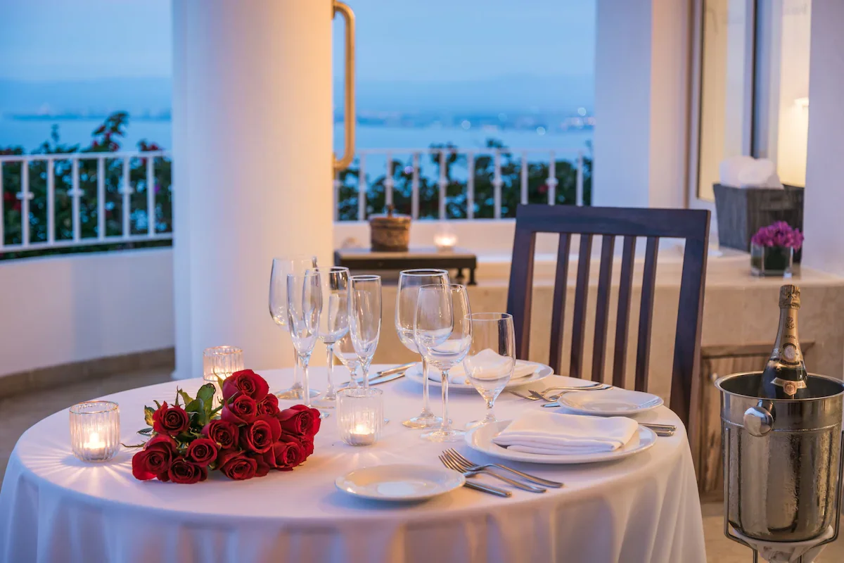 Romantic dinner setup with a white tablecloth, a bouquet of red roses, lit candles, empty wine glasses, and a bottle of champagne in an ice bucket on a balcony with a city view at dusk.