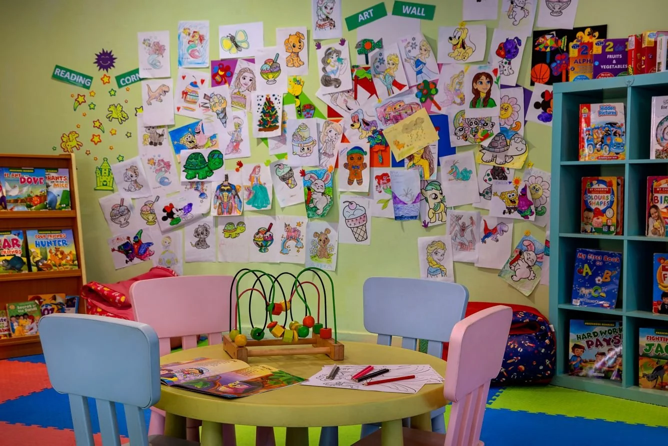 Colorful children's reading corner with a round table, chairs, and walls decorated with assorted hand-drawn and colored pictures of cartoon characters including animals and fairy tale figures. There are bookshelves filled with children's books and an