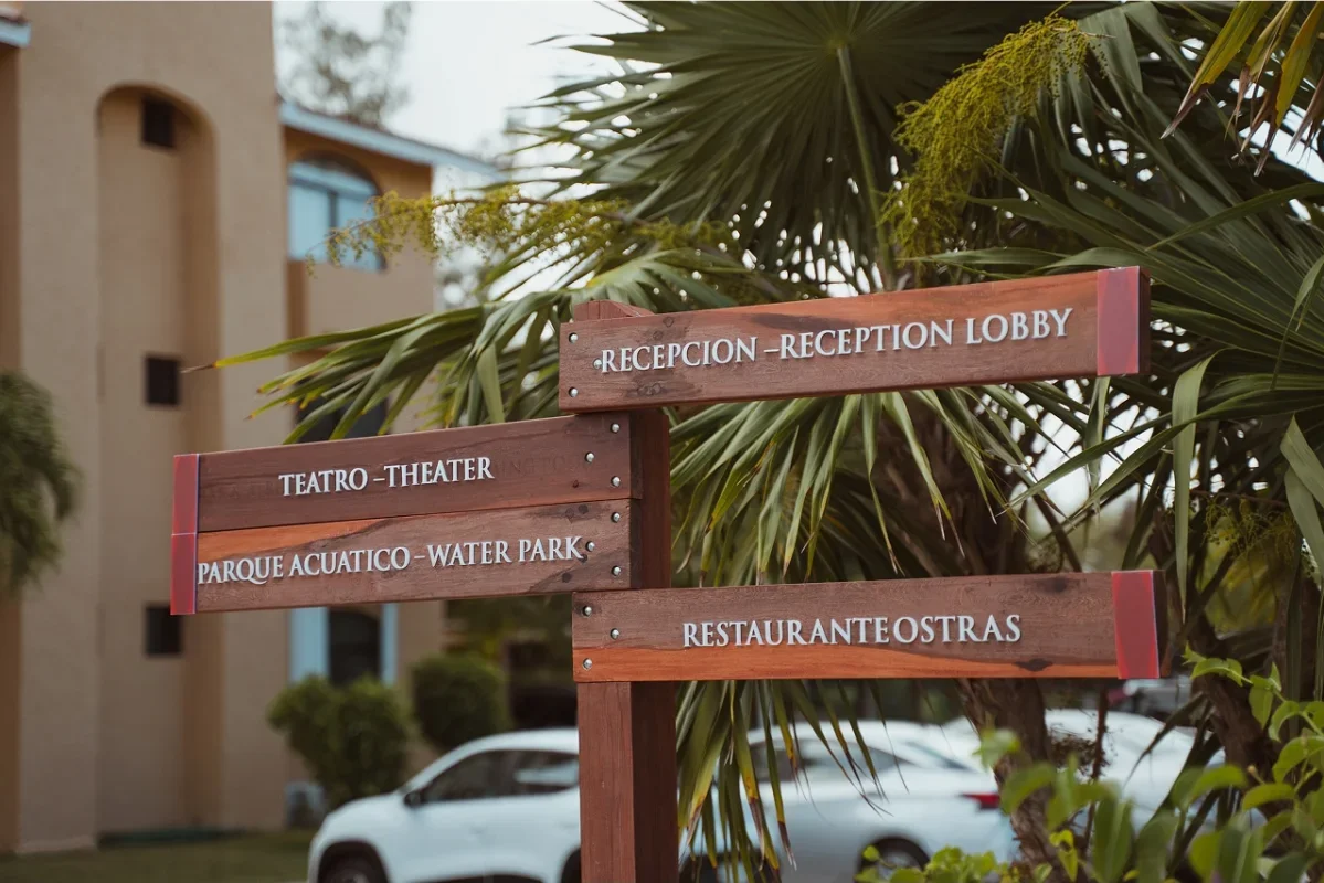 Wooden directional signs with white lettering pointing to the reception, theater, water park, and restaurants, outdoors near tropical plants and trees, with a building and parked cars in the background.