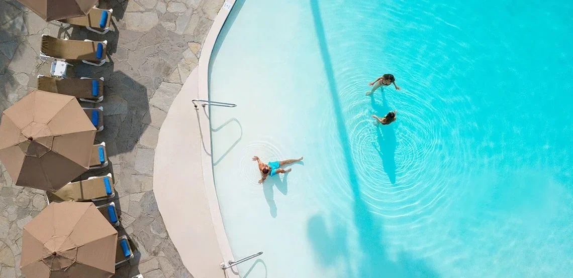 Top-down view of three women swimming and floating in a pool with blue water, surrounded by lounge chairs and umbrellas on a stone pool deck.