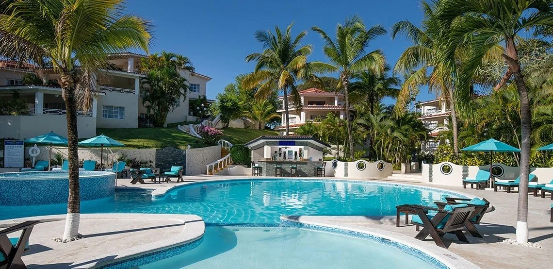 Resort-style swimming pool area with palm trees, lounge chairs, umbrellas, and surrounding residential buildings in a tropical setting.