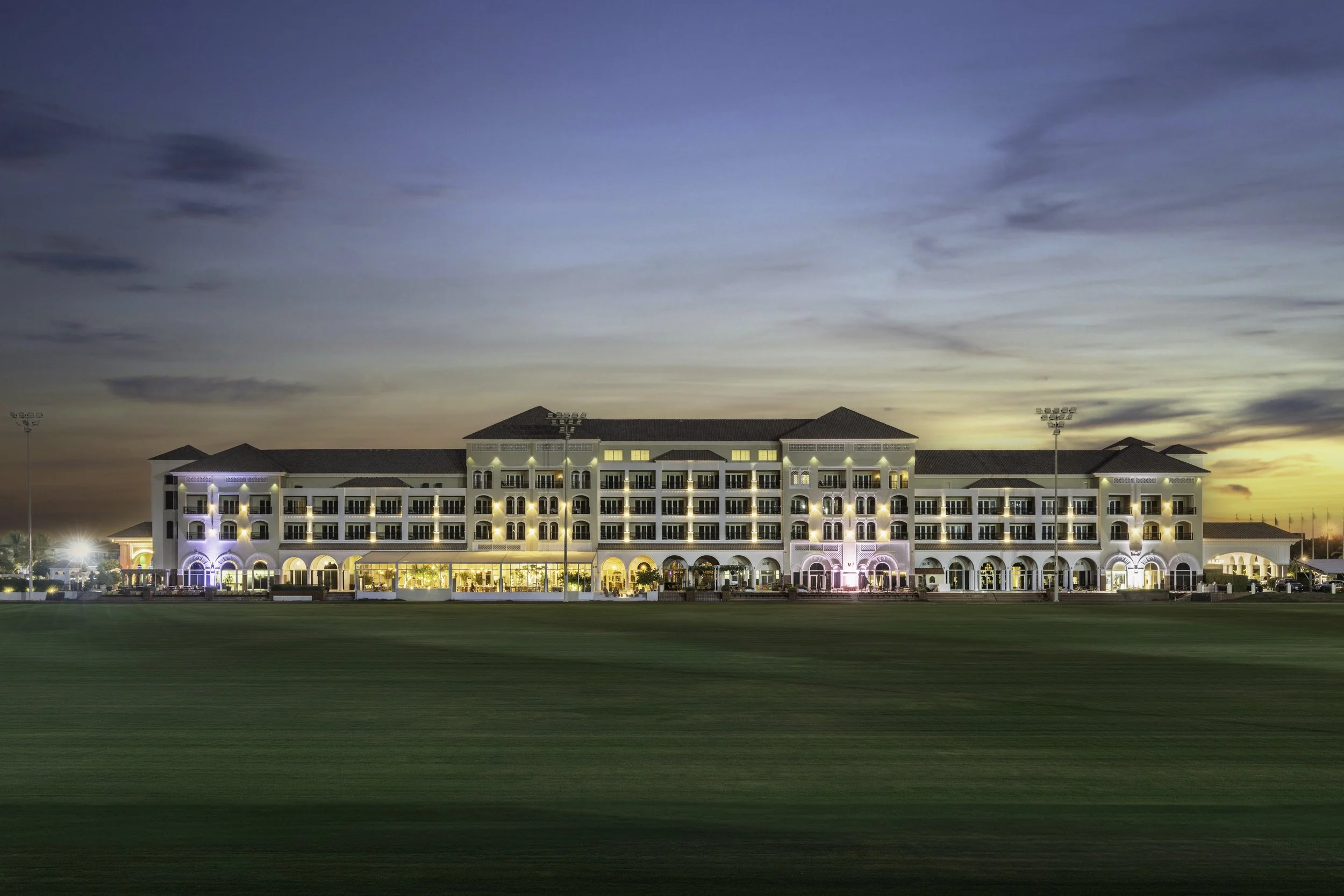 A large hotel or resort building with multiple stories, illuminated with lights, situated on a grassy field during sunset or dusk. The sky is partly cloudy with shades of blue and orange.