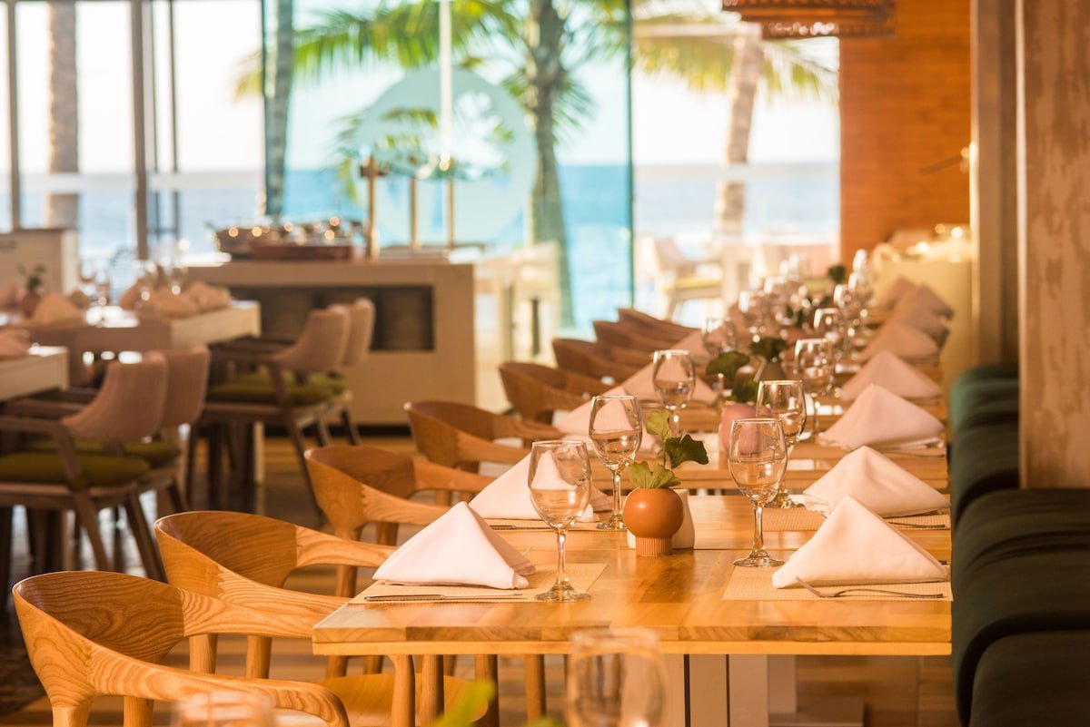 A restaurant dining area with a long wooden table set with wine glasses, white napkins, and small potted plants. There are chairs on one side of the table and a view of the beach with palm trees outside through large windows.